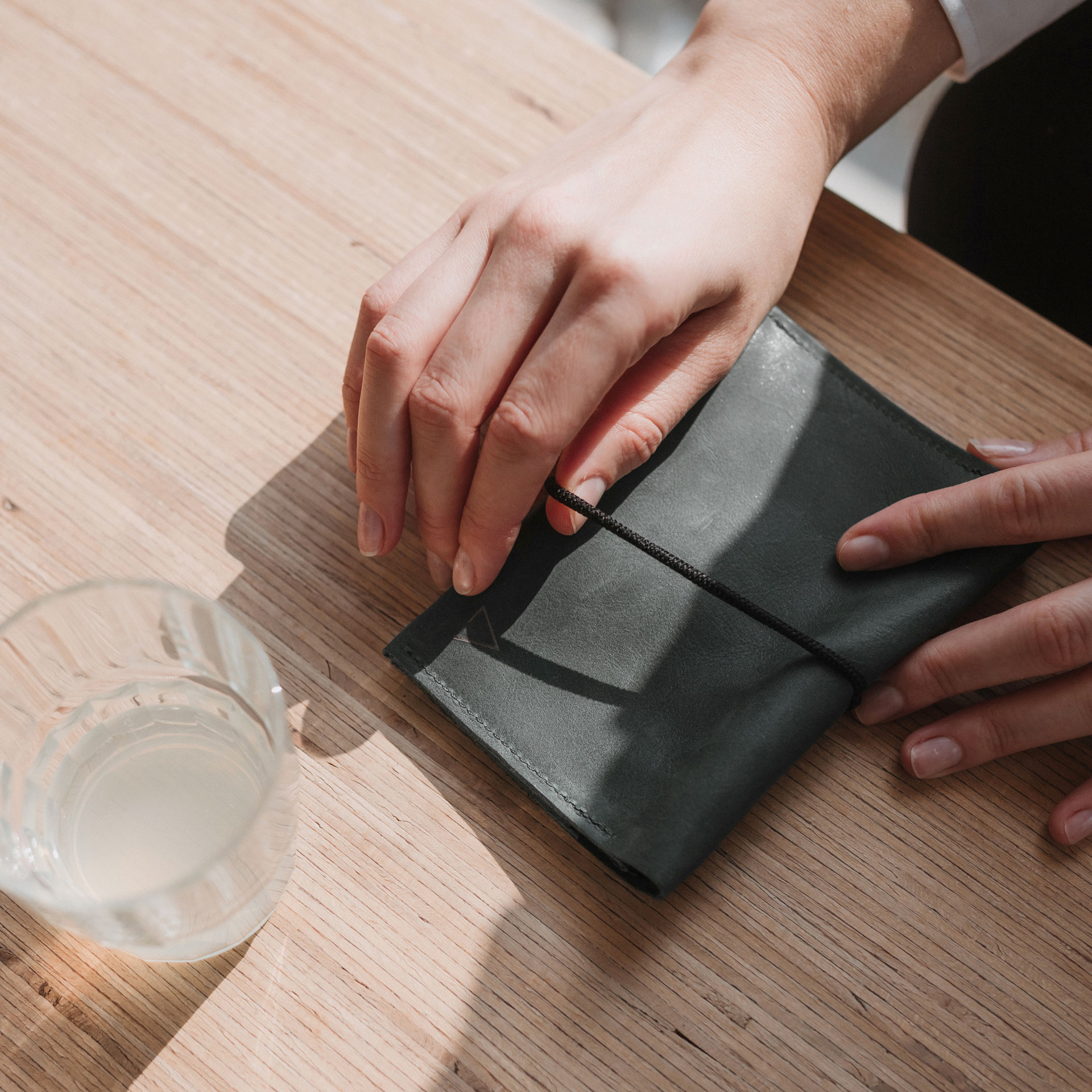 Portemonnaie OLI LARGE in Steingrau mit schwarzem Verschlussband auf Holztisch mit Wasserglas