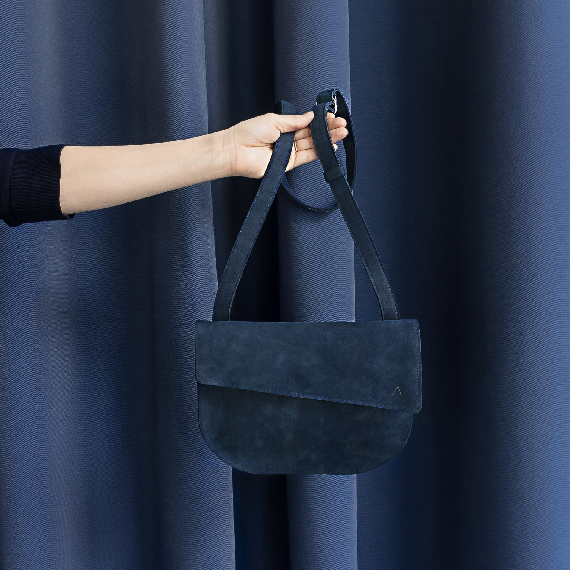 Frau hält Umhängetasche BEA mit verstellbarem Träger aus Naturleder in Blau vor blauem Vorhang