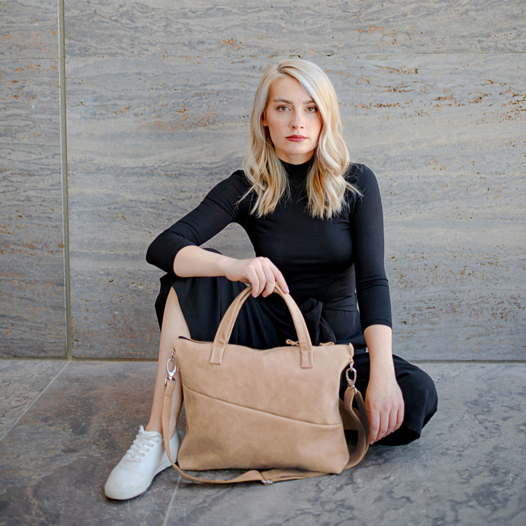 Model sitzt auf dem Boden. Vor ihr steht die Umhängetasche ANA in der Farbe Hellbraun.