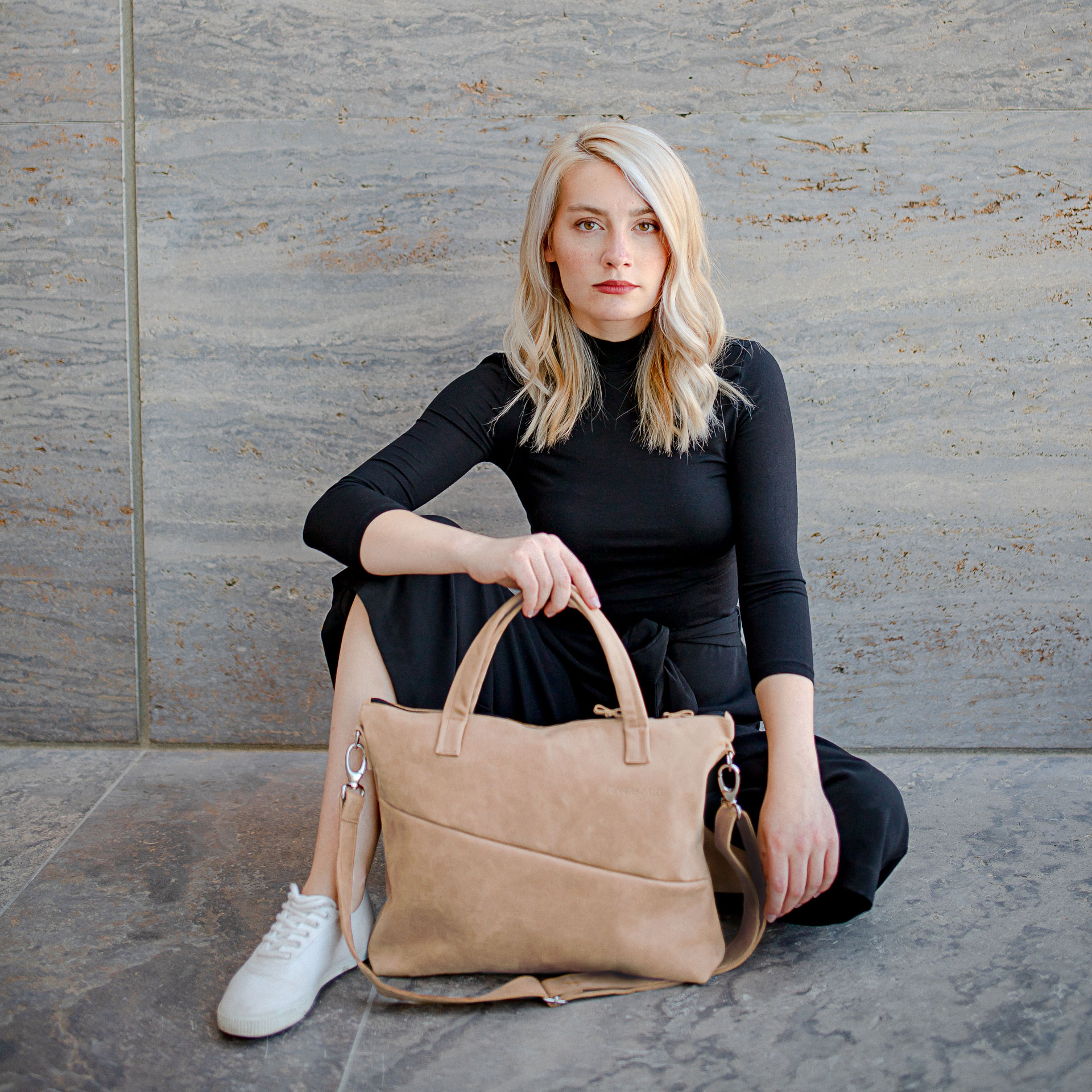Model sitzt auf dem Boden. Vor ihr steht die Umhängetasche ANA in der Farbe Hellbraun.